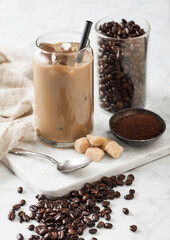 Iced cold caramel coffee with milk and raw beans and spoon with ground coffee and cane sugar on marble board.
