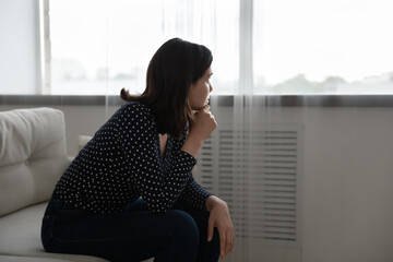 Thoughtful asian woman sit on couch lean forward touch chin look at distance ponder make difficult choice. Pensive young female feel unhappy look aside try to keep emotions under control. Copy space