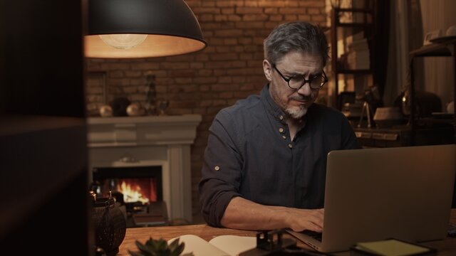 Home office - older white man working from home. Thinking, typing on laptop computer. Sitting at desk in dark living room iin front of fireplace.