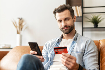 Bearded handsome man makes purchase online. A hipster wearing casual shirt holds a mobile phone and...