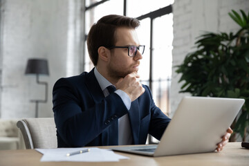 Thoughtful young Caucasian businessman in suit work on computer in office look in distance pondering considering. Pensive male boss or CEO thinking planning, make business decision using laptop.