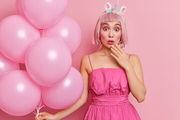 Surprised teenage girl with pink hair stares shocked at camera wears dress stares surprisingly at camera holds bunch of inflated balloons shocked as many guests came on party accepts congratulations
