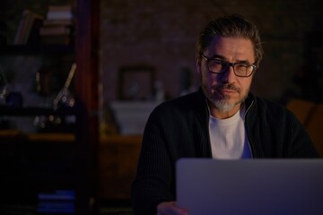 Older man working online with laptop computer at home sitting at desk. Home office, browsing internet, study room. Portrait of mature age, middle age, mid adult man in 50s.