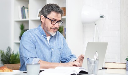 Older man working online with laptop computer at home sitting at desk. Home office, browsing internet, study room. Portrait of mature age, middle age, mid adult man in 50s.