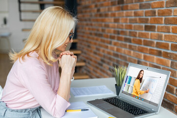 Diligent middle-aged blonde woman sitting at the laptop,listening to the online English lesson,...