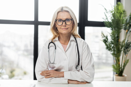 Beautiful female doctor with blond hair and black glasses,wearing lab coat,with a stethoscope,sitting at the desk,smiling,listening the patient,posing for a photo on the clinic site,crossed her arms
