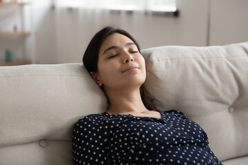 Happy young asian woman lying on couch lean back on pillows nap sleep daydreaming with light smile...