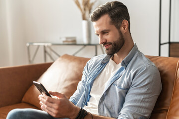 Smiling guy wearing casual outfit messaging, texting on the smartphone. Side view a young man sits on the modern sofa and using mobile app, scrolling feed news, websurfing, playing game