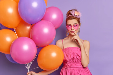 Thoughtful dreamy young woman with hairstyle dressed in festive clothes heart shaped sunglasses celebrates anniversary holds bunch of inflated colorful balloons isolated over purple background