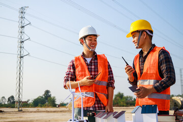 Asian engineer, foreman or leader discussion about home solar cell and wind turbine model in construction site project and High voltage power line pylon in the background. Teamwork, Leadership concept