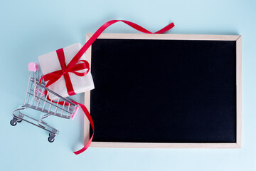 Gift box in shopping carts and blank chalkboard with copy space on light blue background. Seasonal Sale, Discounts, Black Friday concept. Online shopping concept.