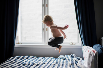 Shirtless boy jumping on bed by window at home