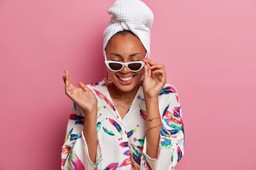 Positive happy woman with dark skin wears sunglasses dressing gown and wrapped towel on head keeps hand raised has fun on domestic party isolated over pink background. People and style concept