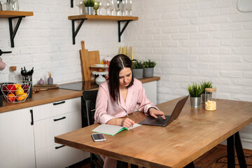 A woman works in the office remotely from home in the kitchen, sitting at the computer. Distance learning online education and work. Girl student prepares for exams.
