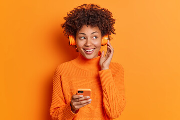 Portrait of good looking African American woman with curly hair smiles broadly has perfect white teeth wears stereo headphones listens favorite music holds smartphone isolated on orange wall