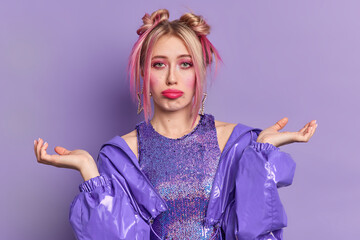 Photo of clueless young European woman looks sadly has two hair buns spreads palms faces difficult choice wears bright vivid makeup dressed in fashionable clothes isolated over purple background