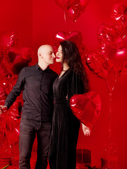 Bald man and young woman in black stand with heart-shaped balloons on red background