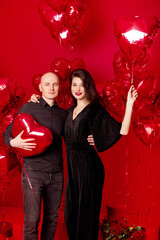 Bald man and young woman in black stand with heart-shaped balloons on red background