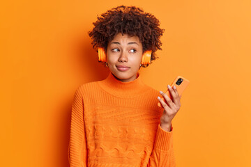 Thoughtful female meloman listens audio track from playlist holds modern smartphone looks pensively...