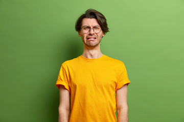 Unhappy handsome Caucasian man cries out in despair feels desperate smirks face wears caual yellow t shirt has melancholic face expression suffers from stress isolated over green background.