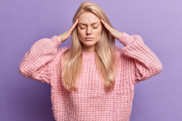 Photo of displeased young blonde woman suffers migraine keeps hands on temples closes eyes suffers...