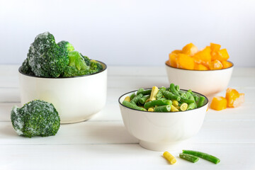 Frozen vegetables assortment. Frozen broccoli, French beans and pumpkin in white bowls on white table