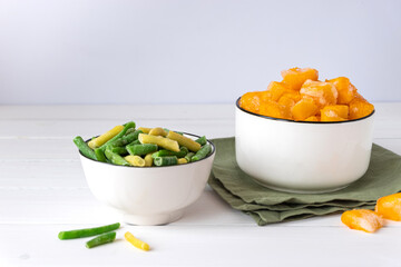 Frozen vegetables assortment. Frozen French beans and pieces of pumpkin in white bowls on white table