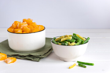 Frozen vegetables assortment. Frozen French beans and pieces of pumpkin in white bowls on white table