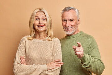 Portrait of happy aged woman and man stand closely to each other dressed in casual jumpers look with great interest in front isolated over brown studio background. People age and emotions concept