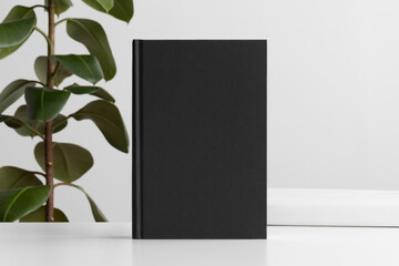 Black book mockup with workspace accessories on the white table and a ficus plant.