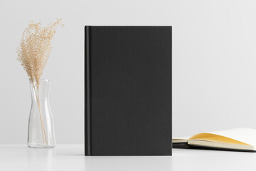 Black book mockup with a dried grass decoration and workspace accessories on the white table.