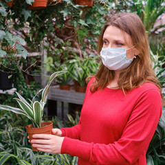 Young woman gardener in medical mask chooses dracaena plant in store. Home gardening hobby during corona virus quarantine