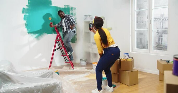 Rear of loving African American wife standing in room taking photos on smartphone of her young husband posing painting wall in green color and smiling. Home repair, house renovation, reconstruction