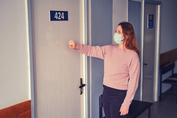 Fototapeta na wymiar A woman in a face mask knocks on the door of the medical office in the corridor hospital. The problem of receiving patients in the clinic during coronavirus