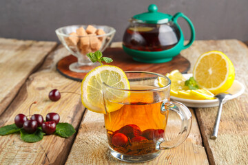 black tea in a glass cup with mint cherries and lemon on a wooden table next to fresh cherries and a teapot and lemon in a plate.