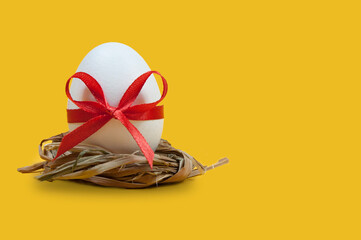white easter egg tied with a red ribbon with a bow in a nest of straw on a yellow background, isolated