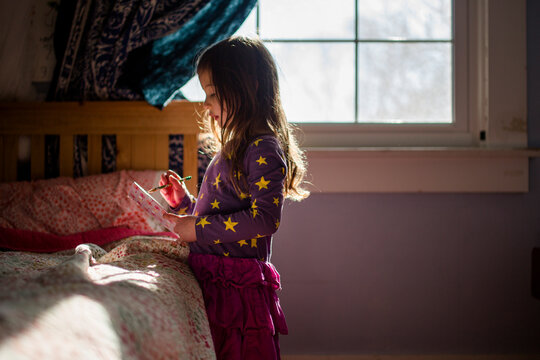 A small girl stands in patch of light in bedroom writing in journal