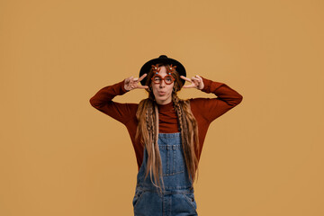 Photo of attractive female farmer looks happy, wears funny glasses. White woman wears denim overall and hat isolated brown color background