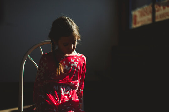 A beautiful little girl sits on a chair in patch of light in dark room