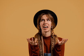 Photo of cute female farmer looks disappointed. White woman wears denim overall and hat isolated brown color background