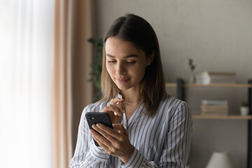 Thoughtful teen female looking on phone screen read pleasant message from beloved person with light smile ponder on text of answer. Interested young lady check mail client on cell wait for good news