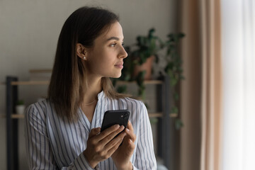Pensive lady stand indoors hold cell look aside relax dream think of positive things. Thoughtful...