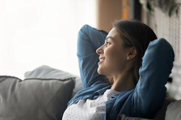 Captured by dreams. Happy serene millennial woman relax on sofa with hands over head look away...