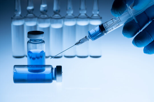 Medical Worker Holds An Ampoule With Vaccine And Medical Disposable Syringe In His Hand. Blue Background. Coronavirus Vaccination Concept Covid 19