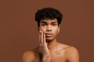 Photo of attractive black man with piercing touches his smooth face and looks at camera. Naked torso, isolated brown color background
