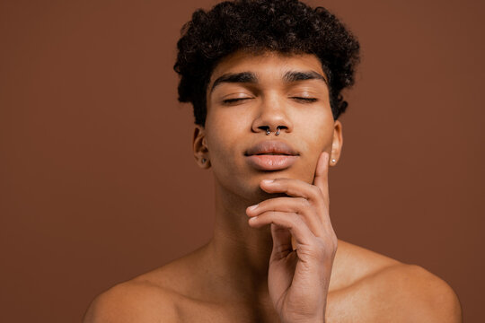 Photo of attractive black man with piercing touches his smooth face with eyes closed. Naked torso, isolated brown color background
