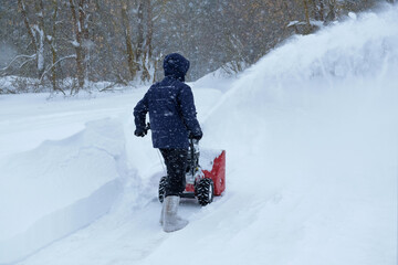 A man working with snowblower to clear driveway near residential house during a heavy snowfall. Russia.