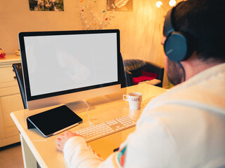 Teleworking - Young man working from his home in his living room, on his computer and his tablet - blank white screen to replace - back view