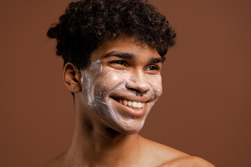 Photo of attractive black man with piercing smiles with nutrition mask on face, nice smile and teeth. Naked torso, isolated brown color background