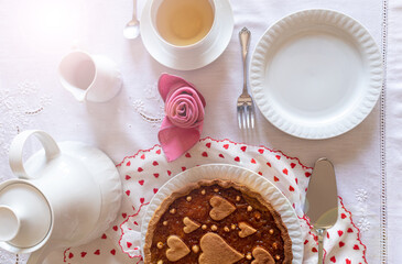 A table set for a special breakfast. An orange tart on a Valentine's fabric is served with a cup of tea near a napkin folded like a rose.